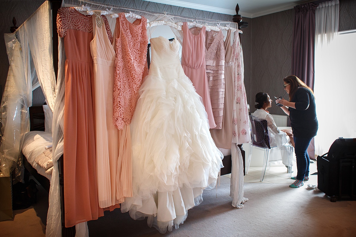 the girls dresses hanging up with the bride having her makeup done in the background