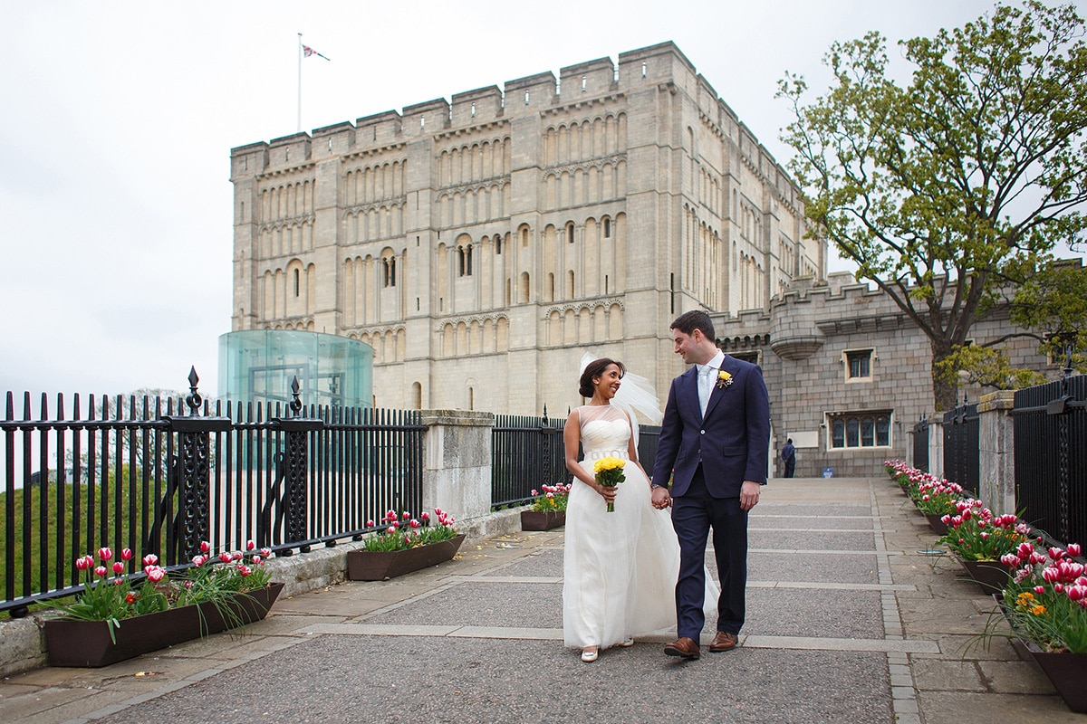 bride and groom walk hand in hand in front of norwich castle