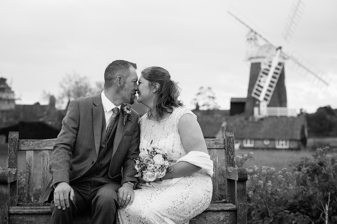 bride and groom on a bench with cley mill in the background